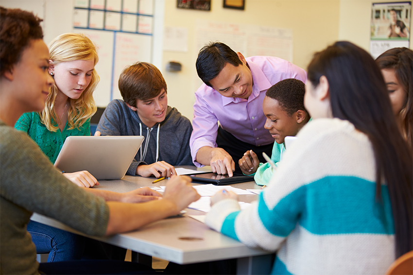 Eine Gruppe Schülerinnen und Schüler unterschiedlicher Herkunft sitzt um einen Tisch und lernt gemeinsam. Ein Lehrer beugt sich über die Schulter eines Schülers und korrigiert die Aufgaben.