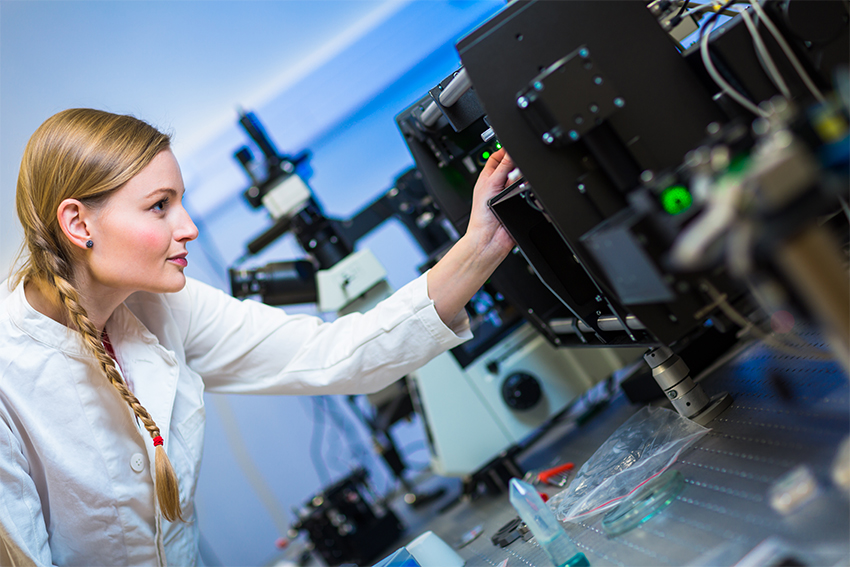 Eine junge blonde Frau in einem Laborkittel beugt sich von links in Bild und justiert ein elektronisches Gerät im rechten Bildteil.