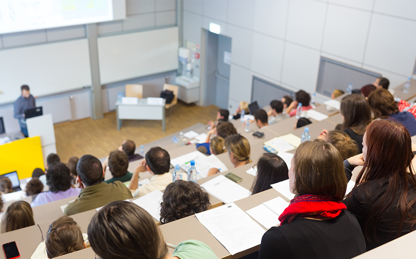 Bild eines Hörsaals mit Studierenden. Zu sehen sind Studierende von hinten in den Hörsaal hinunter fotografiert.