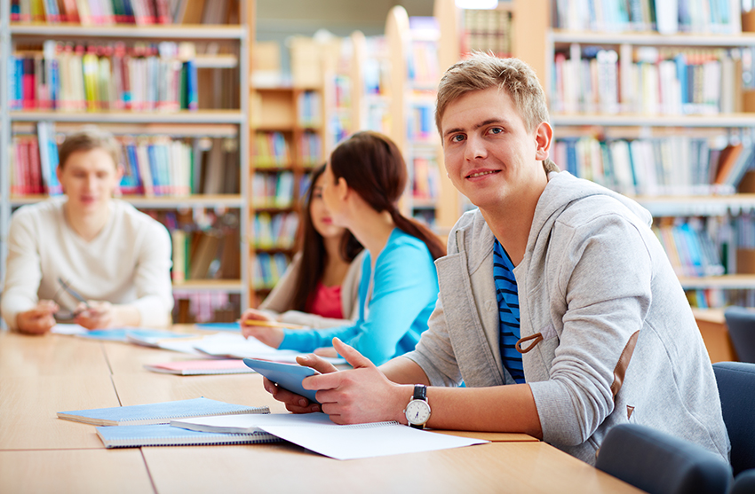 Ein Student sitzt an einem Tisch in einer Bibliothek. Er schaut in die Kamera, im Hintergrund sind weitere Studierende erkennbar.