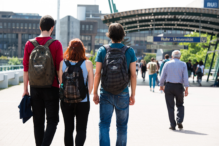 Drei Studierende sind von hinten abgebildet. Sie tragen sommerliche Kleidung und laufen auf der Unibrücke in Richtung der Bahnstation.