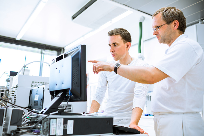 Zwei Männer in weißer Kleidung stehen in einem Labor und schauen gemeinsam auf einen Computerbildschirm.
