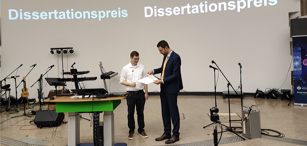 Absolventenfeier2025-Dissertationspreis Dr. Timo Schorlepp is presented with the certificate for the dissertation award at the graduation ceremony. Schorlepp is a dark-haired young man wearing a shirt and glasses.