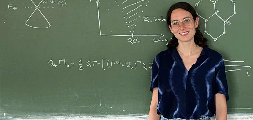 Eine dunkelhaarige junge Frau mit Brille vor einer Tafel, auf der Formeln stehen.