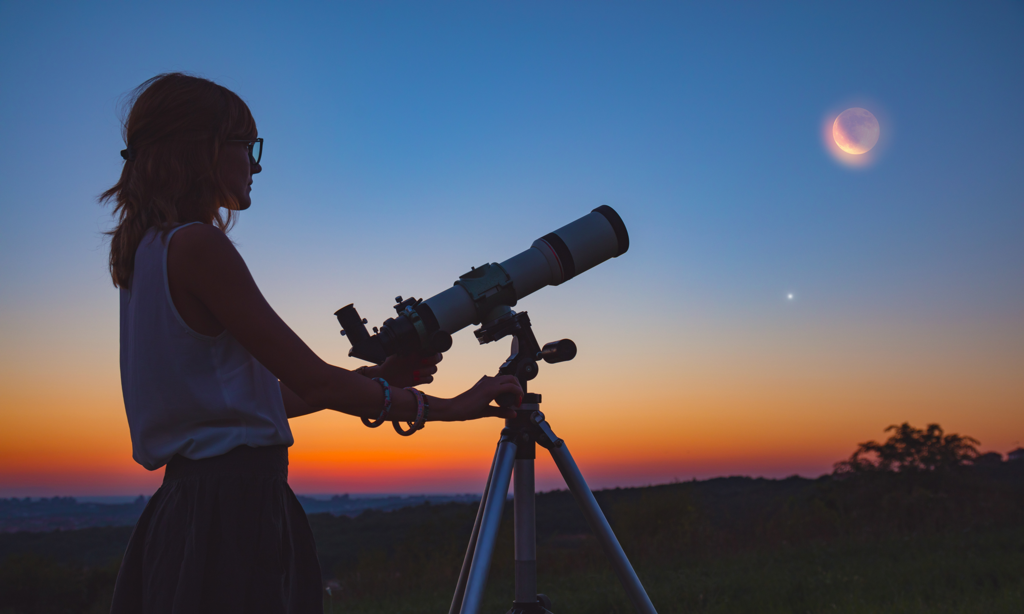 Die Silhouette einer jungen Frau mit Teleskop vor dem Nachthimmel bei Dämmerung