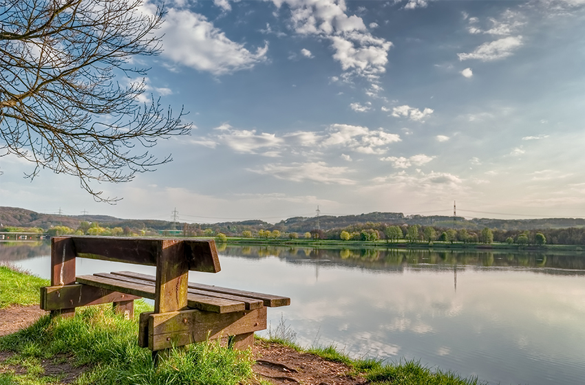 Blick auf den Kemnader See in Bochum. Im linken Vordergrund des Fotos ist eine hölzerne Sitzbank zu sehen.