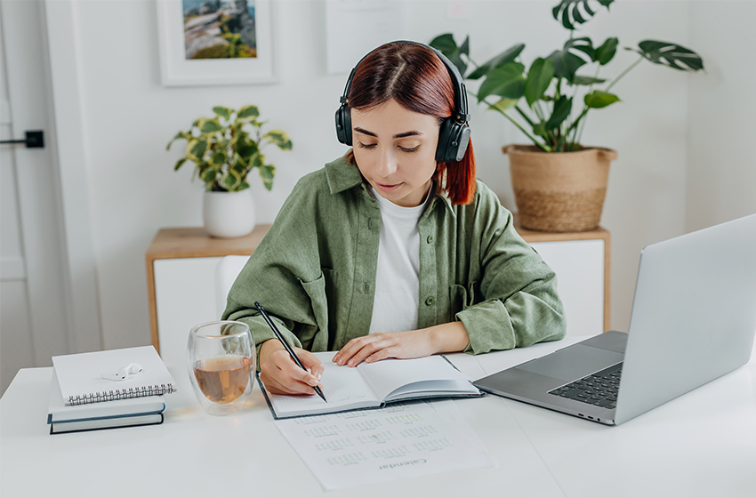 StudentinImHomeoffice Eine Studentin sitzt in ein Notizbuch schreibend vor ihrem geöffnetem Laptop.