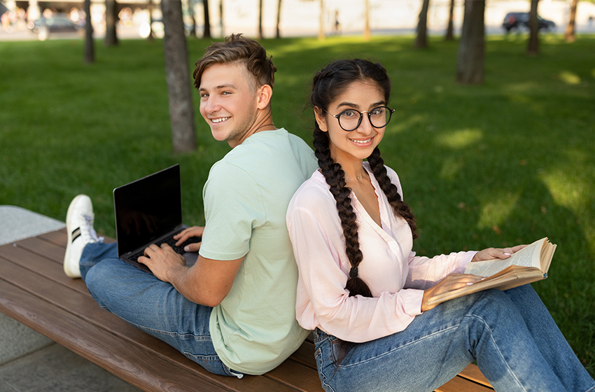 Zwei Studierende sitzen Rücken an Rücken auf einer Bank im Freien und lächeln in die Kamera.