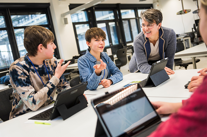 Eine Gruppe Schülerinnen und Schüler unterschiedlicher Herkunft sitzt um einen Tisch und lernt gemeinsam. Ein Lehrer beugt sich über die Schulter eines Schülers und korrigiert die Aufgaben.