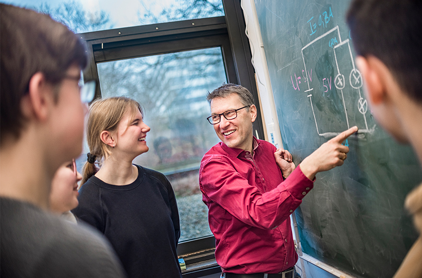 Eine junge Lehrerin mit roten Haaren und einer Brille lächelt in die Kamera. Im Hintergrund kann man einen Klassenraum und eine grüne Tafel erkennen.