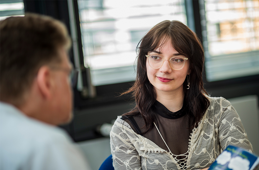 Bild eines Computers und eines Notizbuchs, die auf einem Tisch liegen. Zwei Personen sitzen am Tisch, ihre Gesichter sind nicht erkennbar, eine Person gestikuliert mit ihren Händen