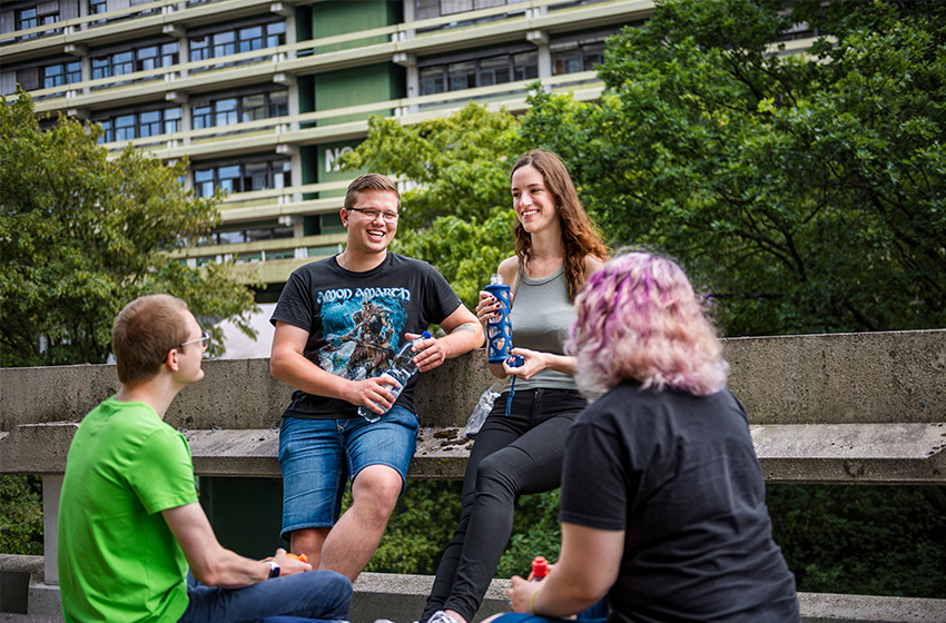 Bild eines Computers und eines Notizbuchs, die auf einem Tisch liegen. Zwei Personen sitzen am Tisch, ihre Gesichter sind nicht erkennbar, eine Person gestikuliert mit ihren Händen