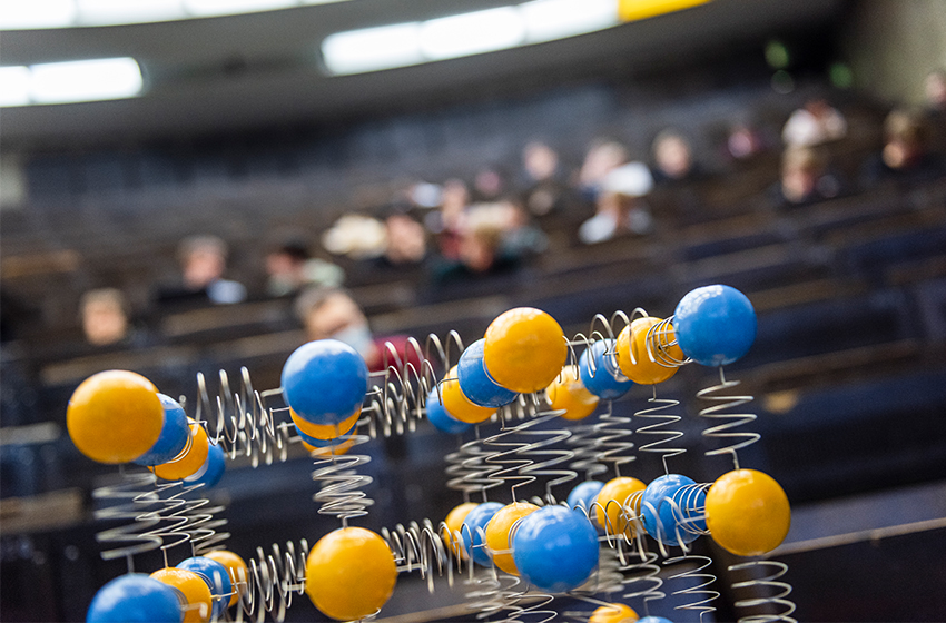 Bild eines Hörsaals mit Studierenden. Zu sehen sind Studierende von hinten in den Hörsaal hinunter fotografiert.